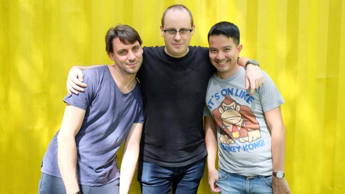 Three men in t-shirts stand before a yellow background in support of Mighty Bear Games releasing Mighty Action Heroes.