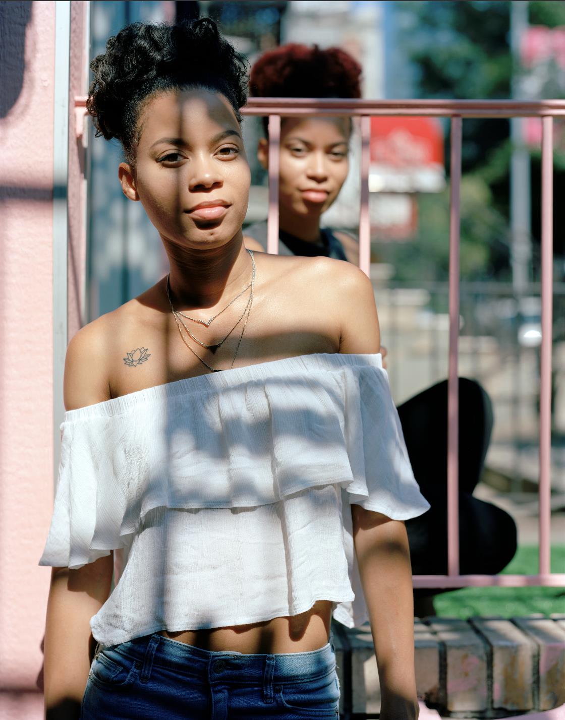 A photograph of two young women who are twins with dark hair and skin standing in a park in a city.