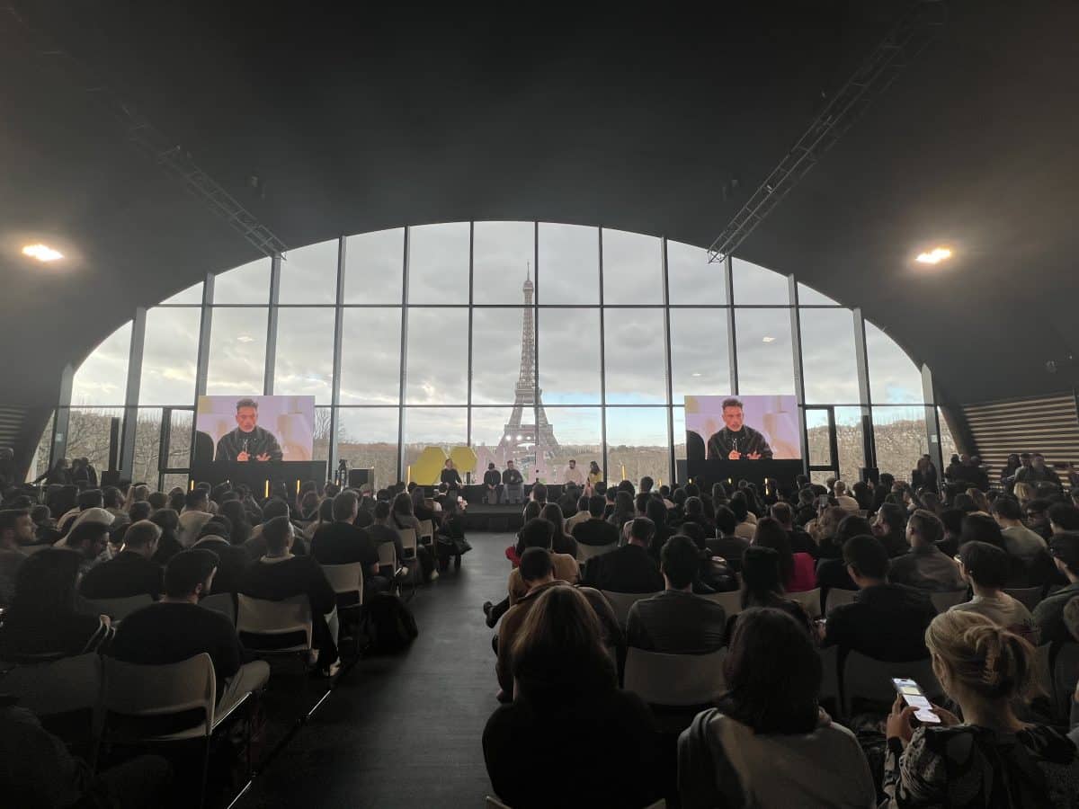 Image of a room full of people listening to speakers with the Eiffel Tower in the background 